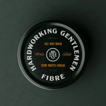 A round black tin of WG Haircare Hardworking Gentlemen Fibre All-Day Hold, shown from a top-down view on a dark charcoal background.